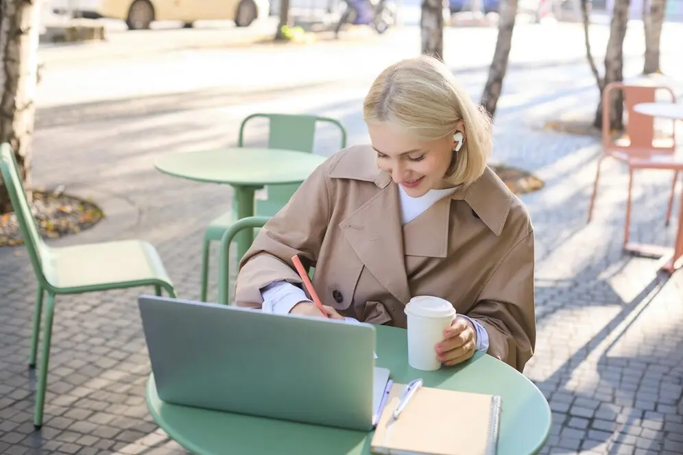 Portrait d'une jeune étudiante blonde dans un café, portant des écouteurs sans fil et travaillant sur son ordinateur portable.
