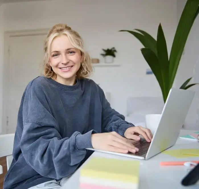Jeune étudiante souriante assise dans sa chambre, faisant ses devoirs à distance sur son bureau.