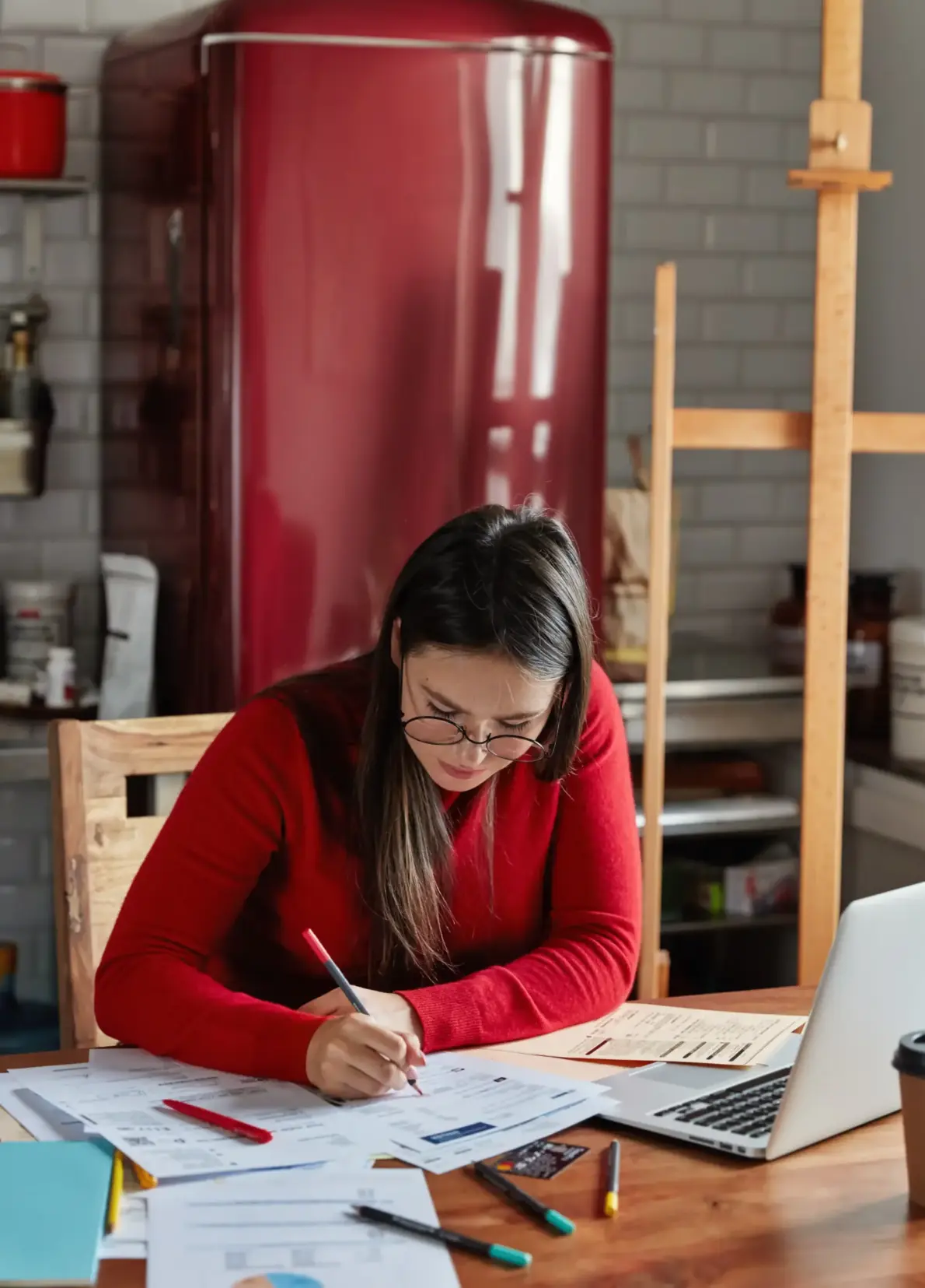 Femme travaillant sur un rapport financier dans une cuisine, avec un réfrigérateur en arrière-plan.