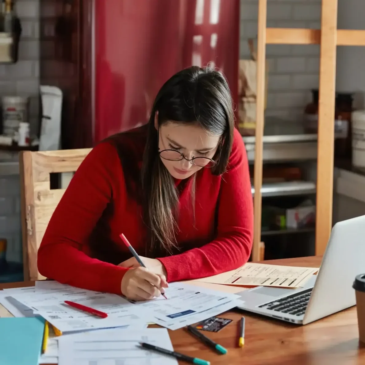 Femme travaillant sur un rapport financier dans une cuisine, avec un réfrigérateur en arrière-plan.