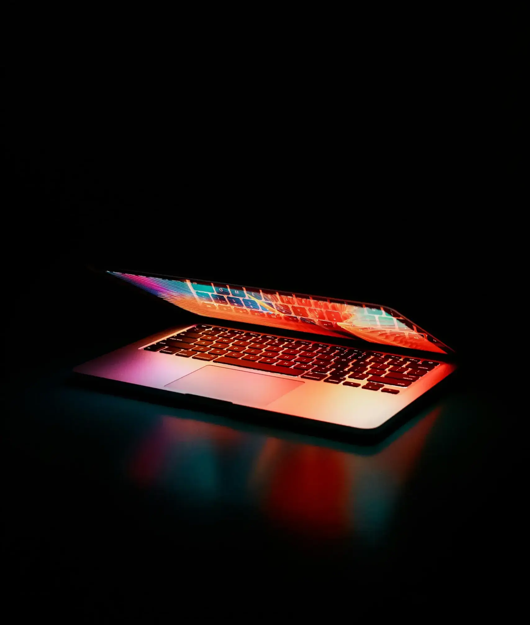 Ordinateur portable partiellement ouvert avec des lumières colorées se reflétant sur le clavier et l'écran, dans une ambiance sombre et moderne.